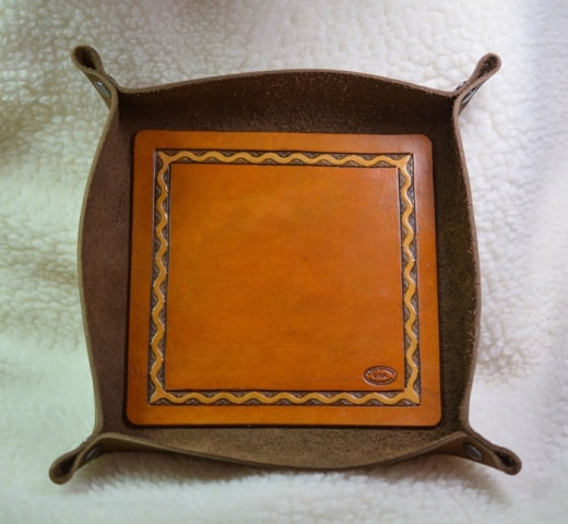 Top view of brown leather tray for keys, change, and other trinkets or as a dice tray, measures about 7-1/2 by 7-1/2 by 1-1/2 inches deep and has a decorative panel with a ribbon border design and smooth center area
